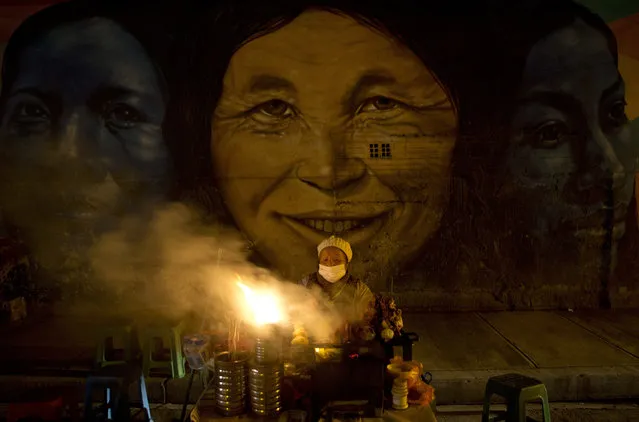 In this October 26, 2018 photo photo, a street vendor keeps food warm on the perimeters of the General Cemetery where images of indigenous women are projected on the wall, in La Paz, Bolivia. People toured the cemetery Friday night for the eighth annual cultural event coined “A night in the cemetery” where actors portray the lives of famous people buried there. (Photo by Juan Karita/AP Photo)