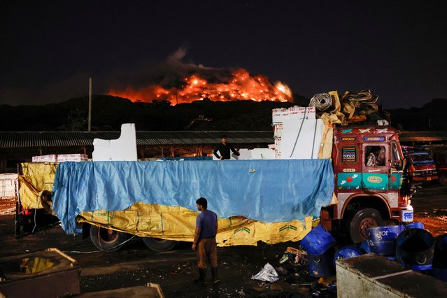 Workers unload boxes containing fish as fire billows from burning garbage at the Ghazipur landfill site, in New Delhi, India on April 22, 2024. (Photo by Adnan Abidi/Reuters)