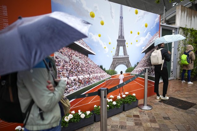 On a day where the weather was different from the illustration on the poster, tennis fans shield themselves from the rain which interrupted the majority of the second round matches of the French Open tennis tournament at the Roland Garros stadium in Paris, Wednesday, May 29, 2024. (Photo by Christophe Ena/AP Photo)