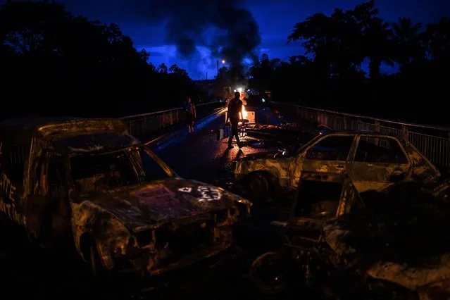 A man carries bottles of water through a road block on fire made of burnt vehicles and debris on his way to the cut-off locality of La Boucan in Sainte-Rose, in the French Caribbean island of Guadeloupe on November 30, 2021, as France's minister for overseas territories left Guadeloupe at an impasse over ways to end more than a week of violent protests sparked by Covid-19 restrictions. Unrest in the former colonial outpost began with a protest over compulsory Covid-19 vaccinations for health workers, but quickly ballooned into a broader revolt over living conditions, and spread to next door Martinique. (Photo by Christophe Archambault/AFP Photo)