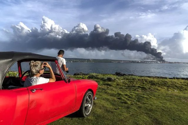People watch a huge plume of smoke rise from the Matanzas supertanker base, as firefighters work to douse a fire that started during a thunderstorm the night before, in Matanzas, Cuba, Sunday, August 7, 2022. Cuban authorities say lightning struck a crude oil storage tank at the base, sparking a fire that sparked four explosions that injured more than 121 people, one person dead and 17 missing. (Photo by Ramon Espinosa/AP Photo)