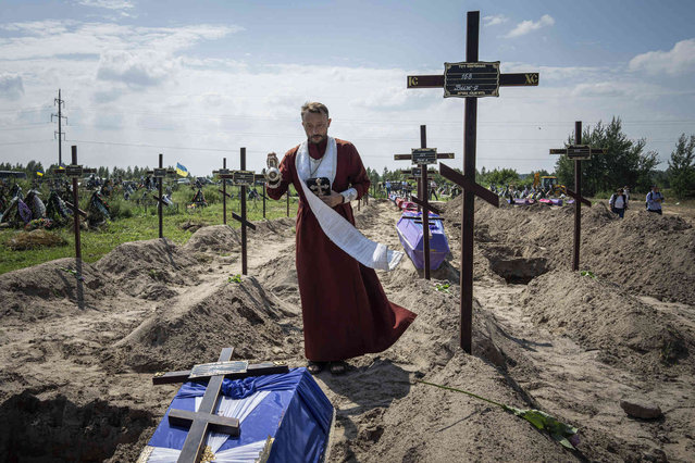 An Orthodox priest, Andrii, prays near the coffins of unidentified civilians killed by Russian troops during Russian occupation in Bucha near Kyiv, Ukraine, on Wednesday, August 17, 2022. Twenty one unidentified bodies exhumed from a mass grave were buried in Bucha on Wednesday. (Photo by Evgeniy Maloletka/AP Photo)