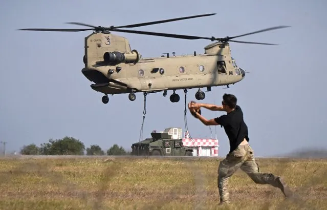 A Boeing CH-47F Chinook tandem rotor helicopter (Vertol) of the US Army's 101st Airborne Division start to lift a military vehicle while in the foreground military personnel play American football during a demonstration drill at Mihail Kogalniceanu Airbase near Constanta, Romania on July 30, 2022. Members of the 101st Airborne Division (Air Assault) headquarters and its 2nd Brigade Combat Team in a ceremony marked their official arrival in Europe at the airbase. The ceremony was followed by a “Romania/US Air and Land Showcase”, a combined US Army and Romanian Army capabilities demonstration. (Photo by Daniel Mihailescu/AFP Photo)