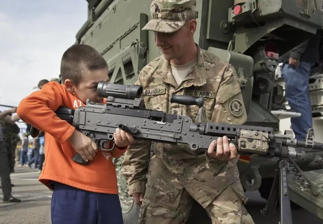 A U.S. Army serviceman helps a Romanian child look through the scope of a machine-gun in Ploiesti, Romania, Wednesday, May 13, 2015. Members of the US military took part in an exercise dubbed the Cavalry March, a drive from southern Romania to a training range in the central region of Transylvania, involving 400 servicemen and 80 armored combat vehicles. (Photo by Vadim Ghirda/AP Photo)