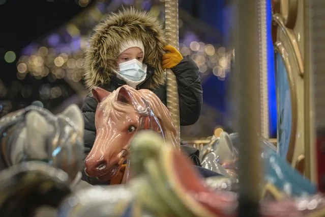 A child wearing a face mask rides in a carousel at a Christmas fair in Bucharest, Romania, Friday, December 17, 2021. The Christmas fairs in the Romanian capital are allowing the public's access to the venues only to the holders of a COVID-19 green pas, proving the owner is fully vaccinated or has recovered after the infection. (Photo by Vadim Ghirda/AP Photo)