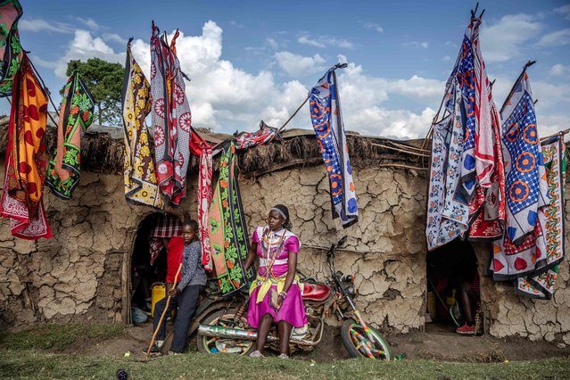 A Maasai woman sits on a motorbike parked next to ceremonial houses decorated with colorful flags at the ceremonial site during the Eunoto ceremony in a remote area near Kilgoris, Kenya on August 18, 2023. Hundreds of young Maasai take part in the Eunoto ceremony, a rite of passage that marks the transition from Moran (young warrior) to adulthood as junior elders. This ceremony is held by every clan once in a generation -every 8 to 10 years- and marks a new age-set. Eunoto, Enkipataa and Olng'esherr are the three main Maasai rites of passage that have been inscribed since 2018 on the Urgent Safeguardiang List of Intangible Cultural Heritage by UNESCO. (Photo by Luis Tato/AFP Photo)