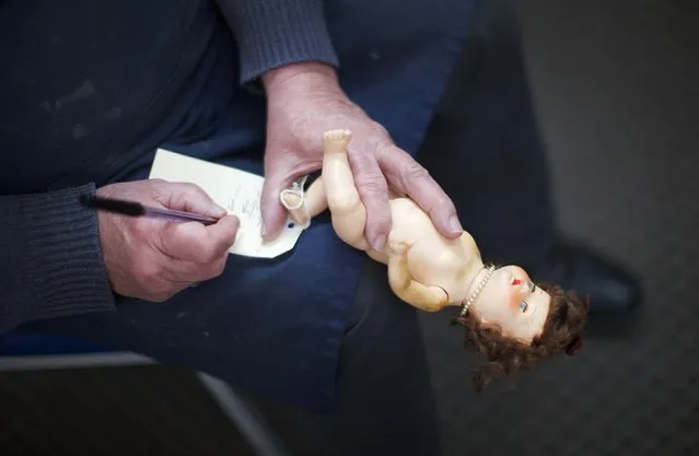 Geoff Chapman, “Head Surgeon” and third-generation owner of Sydney's Doll Hospital, holds a small doll on one hand as he writes details onto its repair card attached to its foot, at his workshop June 28, 2014. (Photo by Jason Reed/Reuters)