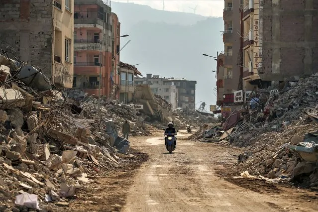 A man rides a motorcycle past debris from destroyed buildings in Samandag, southern Turkey, Wednesday, February 22, 2023. Survivors of the earthquake that jolted Turkey and Syria 15 days ago, killing tens of thousands of people and leaving hundreds of thousands of others homeless, dealt with more trauma and loss Tuesday after another deadly quake and aftershocks rocked the region. (Photo by Emrah Gurel/AP Photo)