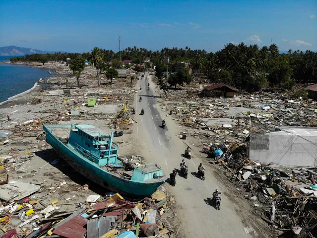 People drive past a washed up boat and collapsed buildings in Palu on October 1, 2018, after an earthquake and tsunami hit the area on September 28. Mass graves were being readied on October 1 for hundreds of victims of the Indonesian quake and tsunami as authorities battled to stave off disease and reach desperate people still trapped under shattered buildings. (Photo by Jewel Samad/AFP Photo)
