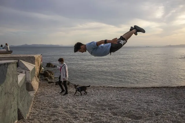 25 year-old Jimmakos Scotis makes a backflip as a woman wearing a protective face mask walks with her dog at the seaside of Alimos, suburb of Athens, on Tuesday, May 4, 2021. Greece began easing coronavirus-related restrictions with a view to opening to the vital tourism industry in the summer. (Photo by Petros Giannakouris/AP Photo)