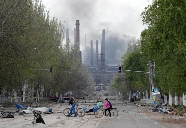 People walk their bikes across the street as smoke rises above a plant of Azovstal Iron and Steel Works during Ukraine-Russia conflict in the southern port city of Mariupol, Ukraine on May 2, 2022. (Photo by Alexander Ermochenko/Reuters)