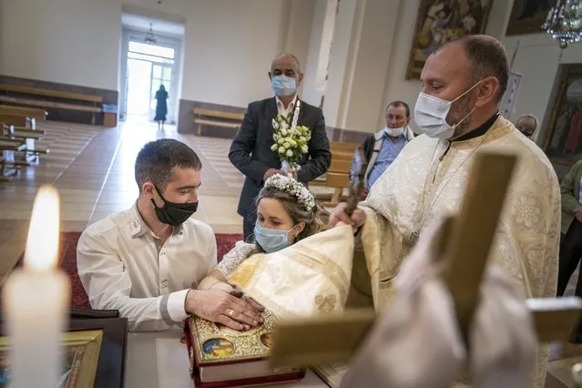 In this photo taken on Sunday, May 24, 2020, Father Vasyl Gasynets, Ukrainian Greek Catholic Church priest, right, conducts a religion service while Anatoliy Megeden and Svetlana Megeden swearing on the Holy Bible during their wedding ceremony at a church in Chernivtsi. Gasynets has returned to conducting services at his Greek Catholic church in priestly raiment, but wearing a mask while distributing communion. (Photo by Evgeniy Maloletka/AP Photo)