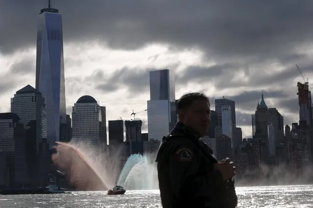 A Jersey City Fire Department boat throws water with the colors red, blue, and white along the Hudson river while a New Jersey police officer stands guard during a ceremony for 9/11 victims at a memorial, across from New York's Lower Manhattan and One World Trade Center in Exchange Place, New Jersey September 11, 2015. (Photo by Eduardo Munoz/Reuters)