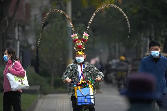 A man wearing a face mask dressed with Monkey King costume headgear rides past masked residents on a street in Beijing, Wednesday, November 2, 2022. (Photo by Andy Wong/AP Photo)