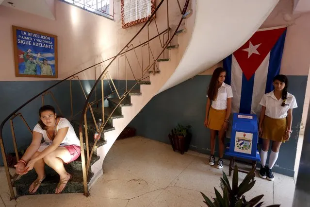 A young woman sits on a staircase as two school girls stand next to a ballot box at a polling station at a school during the municipal elections in Havana April 19, 2015. The writing on the poster showing Cuba's former President Fidel Castro (L) and his brother Cuba's President Raul Castro reads, “The revolution, powerful and victorious, goes ahead”. (Photo by Reuters/Stringer)