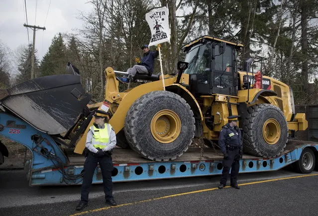 Royal Canadian Mounted Police officers stand by as a man sits with his leg locked to a piece of heavy equipment being delivered to Kinder Morgan in Burnaby, British Columbia, Monday, March 19, 2018. Protesters who blockaded an entrance - defying a court order - were arrested earlier in the day while protesting the Kinder Morgan Trans Mountain pipeline expansion. (Photo by Darryl Dyck/The Canadian Press via AP Photo)