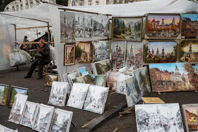 A street vendor selling paintings reads a newspaper while waiting for customers on a sidewalk in central Lviv, Ukraine, November 25, 2016. (Photo by Gleb Garanich/Reuters)