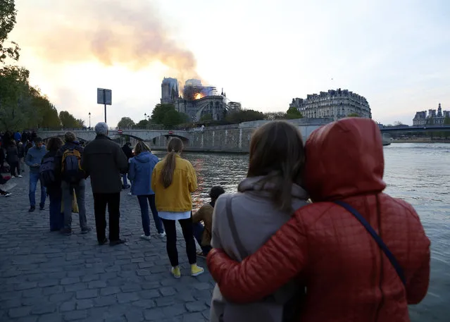 People watch as flames and smoke rise from Notre Dame cathedral as it burns in Paris, Monday, April 15, 2019. Massive plumes of yellow brown smoke is filling the air above Notre Dame Cathedral and ash is falling on tourists and others around the island that marks the center of Paris. (Photo by Thibault Camus/AP Photo)