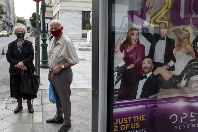 Two people wearing face masks to prevent the spread of COVID-19, wait at a bus station where an advertisement for a TV show is displayed, in Athens, Monday, November 2, 2020. The government announced new lockdown measures Saturday to stem the rapid rise in new cases, among which are the closure of bars, cafes, restaurants and gyms in large swaths of the country, will take effect Tuesday through at least the end of November. (Photo by Yorgos Karahalis/AP Photo)
