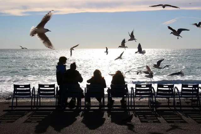 Seagulls fly over tourists looking out over the Mediterranean sea, on the Promenade Des Anglais in Nice, southeastern France, Friday, January 8, 2016. Temperatures in the area rose to 18 degrees Celsius (64 Fahrenheit). (Photo by Lionel Cironneau/AP Photo)
