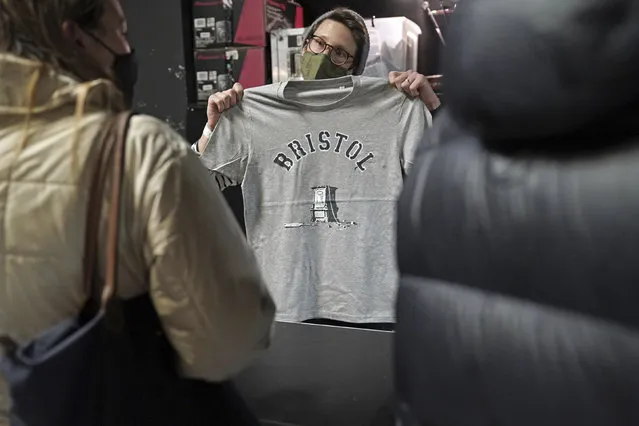 A person inside Rough Trade in Bristol, England, Saturday December 11, 2021, holds up a T-shirt designed by street artist Banksy, being sold to support four people facing trial accused of criminal damage in relation to the toppling of a statue of slave trader Edward Colston. The anonymous artist posted on Instagram pictures of limited edition grey souvenir T-shirts which will go on sale on Saturday in Bristol. The shirts have a picture of Colston's empty plinth with a rope hanging off, with debris and a discarded sign nearby. (Photo by Jacob King/PA Wire via AP Photo)