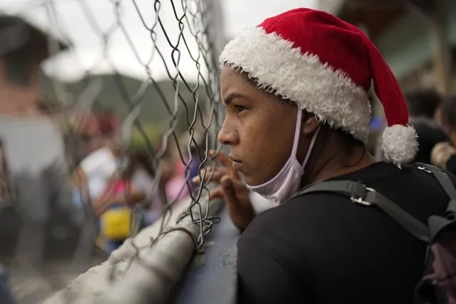 A woman wearing a Santa Claus hat looks from the fence as she waits for the start of a Christmas performance at a square in the Mamera neighborhood of Caracas, Venezuela, Thursday, December 16, 2021. (Photo by Ariana Cubillos/AP Photo)