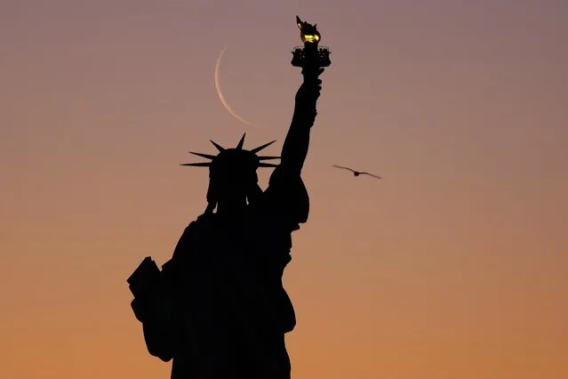 A 4 percent illuminated moon rises behind the Statue of Liberty as the sun rises in New York City on January 30, 2022, as seen from Jersey City, New Jersey. (Photo by Gary Hershorn/Getty Images)