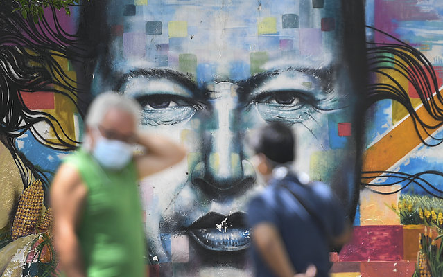 Pedestrians wearing masks to curb the spread of the new coronavirus chat in front of a mural of late President Hugo Chavez, in Caracas, Venezuela, Sunday, July 12, 2020. (Photo by Matias Delacroix/AP Photo)