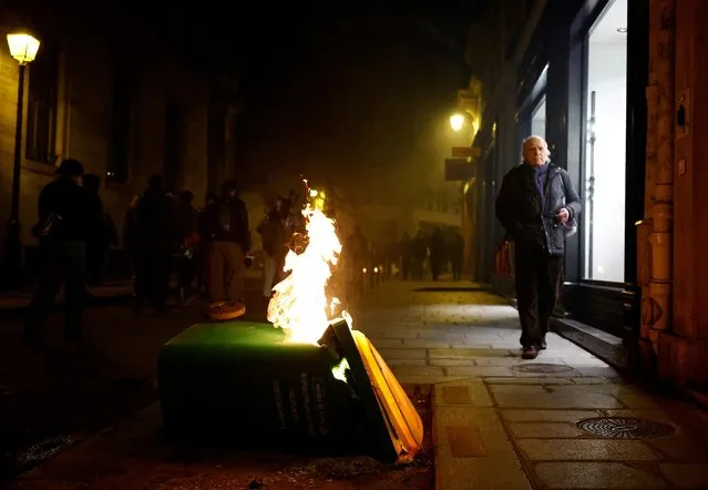 People walk near a trash bin on fire, during a gathering to condemn police violence, and in support of two protesters who fell into a coma after being injured in clashes against police in Sainte-Soline, in Paris, France on March 30, 2023. (Photo by Sarah Meyssonnier/Reuters)