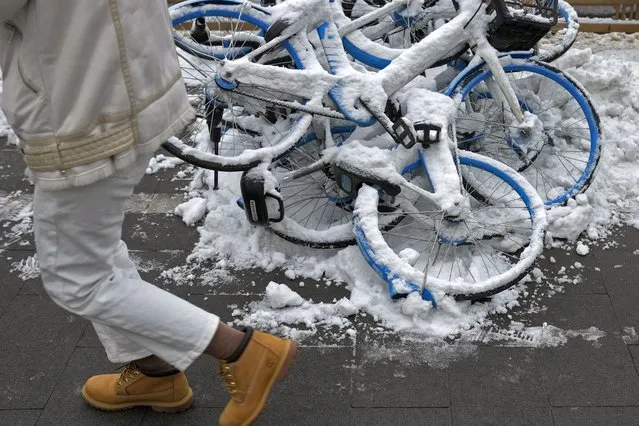 A resident walks by snow covered bicycle of bike-sharing companies piled up on a sidewalk after a snow fall in Beijing, Monday, December 11, 2023. An overnight snowfall across much of northern China prompted road closures and the suspension of classes and train service on Monday. (Photo by Andy Wong/AP Photo)