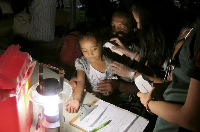 In this October 8, 2015 photo, Keioleen Helly, 3, center, is held by her grandmother Kionina Kaneso as she gets her temperature taken by medical student Ailea Apana at the University of Hawaii John A. Burns School of Medicine in Honolulu. The family was getting shots for a tuberculosis test required for entry to most shelters from volunteers at a mobile medical van by lantern light. (Photo by Cathy Bussewitz/AP Photo)