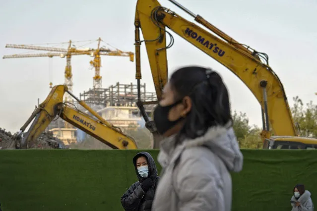 Women wearing face masks to help curb the spread of the coronavirus walk by machinery dismantling buildings during the morning rush hour in Beijing, Tuesday, December 1, 2020. (Photo by Andy Wong/AP Photo)