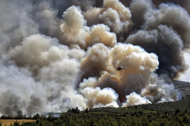 A firefighting helicopter flies in front of a thick cloud of smoke from a forest fire at Spathovouni village, near Corinth, southwest of Athens, Friday, July 23, 2021. Fire brigade vehicles are assisted by two helicopters and four firefighting planes in an effort to extinguish the fire. (Photo by Valerie Gache/AP Photo)