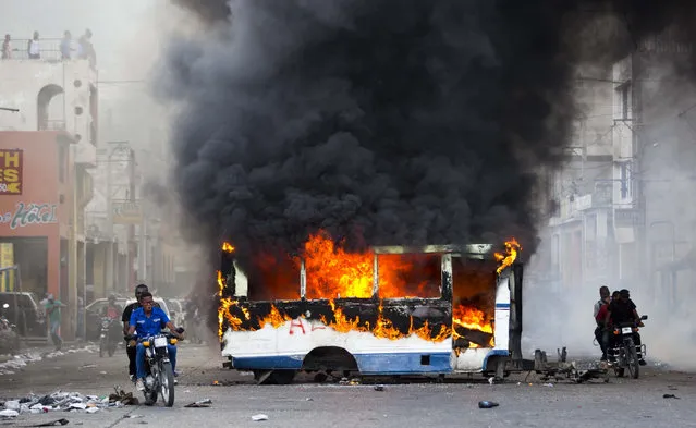 Motorcyclists pass a burning bus, set fire by opposition protesters demanding to know how Petro Caribe funds have been used by the current and past administrations, on the sidelines of events marking the 215th anniversary of independence Battle of Vertieres in Port-au-Prince, Haiti, Sunday, November 18, 2018. Much of the financial support to help Haiti rebuild after the 2010 earthquake comes from Venezuela's Petro Caribe fund, a 2005 pact that gives suppliers below-market financing for oil and is under the control of the central government. (Photo by Dieu Nalio Chery/AP Photo)