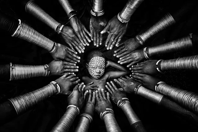 “Tribal Identity”. A young Suri boy, his face painted with white clay, is surrounded by women who have decorated their arms with bronze bracelets. The tribe in the Omo Valley of Ethiopia maintains important symbols of tribal identity, such as face and body painting. The shapes and colors convey strong bonds and meaning. (Photo by Zay Yar Lin/International Portrait Photographer of the Year)