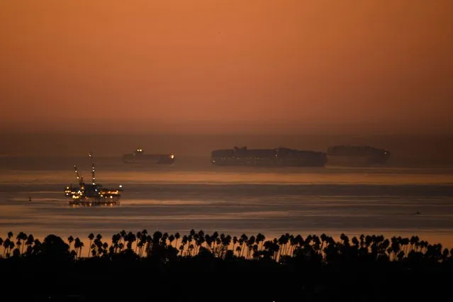 An oil platform (L) stands offshore as cargo shipping container ships wait in the Pacific Ocean to enter the Port of Los Angeles and Port of Long Beach on October 15, 2021 as seen before sunrise from Signal Hill, California. The port, North America's busiest container terminal, began 24-hour operations October 14, 2021 after the White House intervened to help ease bottlenecks that are choking commerce and pushing up prices. (Photo by Patrick T. Fallon/AFP Photo)