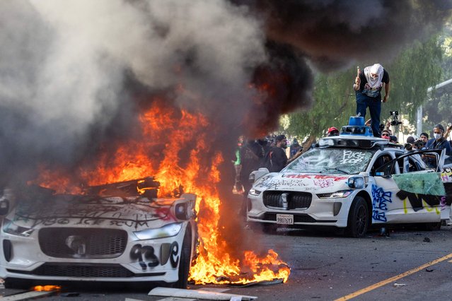 Demonstrators smash the windshield of a vehicle next to a burning Waymo vehicle as protesters clash with law enforcement in the streets surrounding the federal building during a protest following federal immigration operations in Los Angeles, California, on June 8, 2025. Demonstrators torched cars and scuffled with security forces in Los Angeles on June 8, as police kept protestors away from the National Guard troops President Donald Trump sent to the streets of the second biggest US city. (Photo by Ringo Chiu/AFP Photo)
