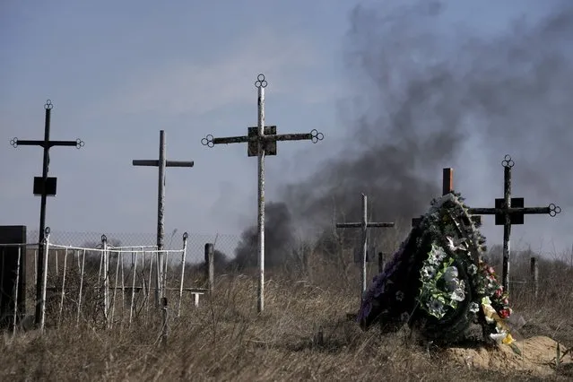 A smoke from shelling rises as a wreath of flowers is placed at a cemetery in Vasylkiv south west of Kyiv, Ukraine, Saturday, March 12, 2022. Russian forces appeared to make progress from northeast Ukraine in their slow fight to reach the capital, Kyiv, while tanks and artillery pounded places already under siege. (Photo by Vadim Ghirda/AP Photo)