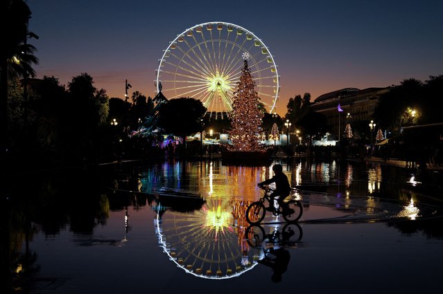 A boy rides a bicycle by a ferris wheel and a Christmas tree set up in the French riviera city of Nice on December 15, 2025. (Photo by Valery Hache/AFP Photo)