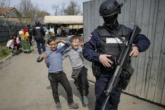 Children play next to an armed riot police officer during a raid of the National Environmental Guard through scrape littered back yards in Vidra, Romania, Tuesday, April 13, 2021. Many people in the Roma community in SIntesti scrape a dangerous living by illegally setting fire to whatever they can find that contains metal, from computers to tires and electrical cables. (Photo by Vadim Ghirda/AP Photo)