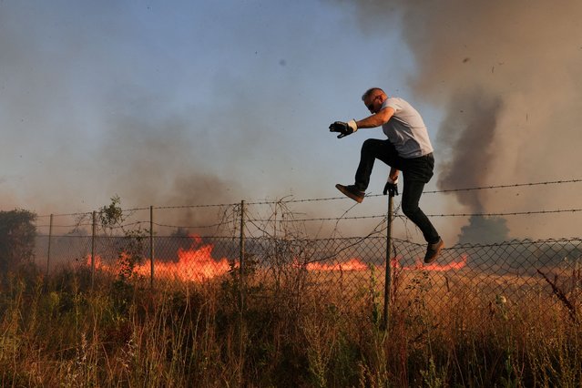 A man jumps as flames and smoke rise from a wildfire in a village in Dubrava, Kosovo, on July 22, 2025. (Photo by Valdrin Xhemaj/Reuters)