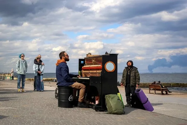 Street musician from Kyiv, Ihor Yanchuk, conducts an impromptu piano concert on the beach to support residents as smoke rises over the port of Pivdennyi after Russian missiles struck, amid Russia's attack on Ukraine, in Odesa, on April 19, 2024. (Photo by Nina Liashonok/Reuters)