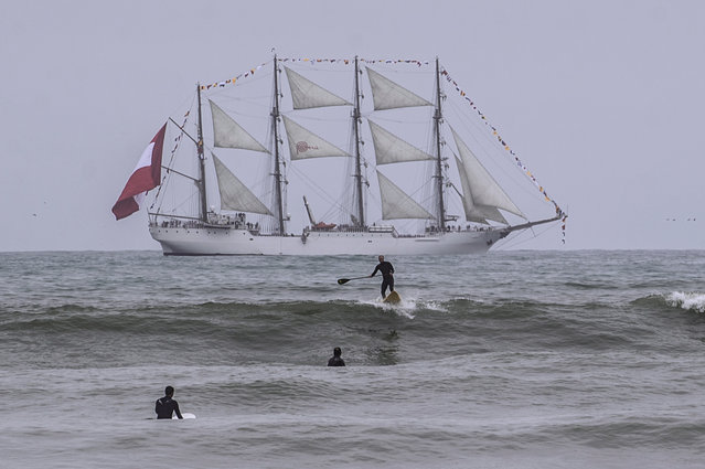 A paddle boarder surfs a wave with Peru's navy flagship “Union” on the background at La Pampilla beach in Costa Verde bay, in Lima, on July 27, 2019. It was the ancient inhabitants of Peru the first to ride waves, surf instructor Elias Panta tells AFP. They did it in narrow reed-made vessels known as “caballitos de totora” (little reed horses). On July 29, 2019, surfing makes it debut as a Pan-American sport in the Lima 2019 Games currently being held in Peru. (Photo by Ernesto Benavides/AFP Photo)