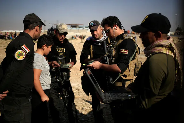 A boy, who just fled the Samah district of eastern Mosul, stands with the Special Forces holding men's identification cards, as they conduct interrogations to ensure the displaced do not belong to the Islamic State group, at the Iraqi Special Forces checkpoint in Kokjali, east of Mosul, Iraq November 5, 2016. (Photo by Zohra Bensemra/Reuters)