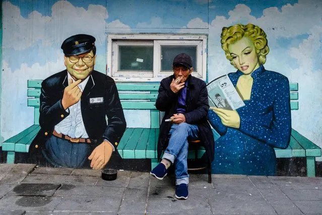 A man smokes a cigarette as he sits in front of a mural depicting South Korean television host Song Hae (L) and US actress Marilyn Monroe (R) outside a cinema in Seoul on January 5, 2023. (Photo by Anthony Wallace/AFP Photo)