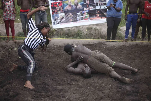 Ugandan youths perform an amateur wrestling tangle in the soft mud in Kampala, Uganda Wednesday, March. 20, 2023. The open-air training sessions, complete with an announcer and a referee, imitate the pro wrestling contests the youth regularly see on television. While a pair tangles inside the ring, made with bamboo poles strung with sisal rope, others standing ringside cheer feints and muscular shows of strength. (Photo by Patrick Onen/AP Photo)