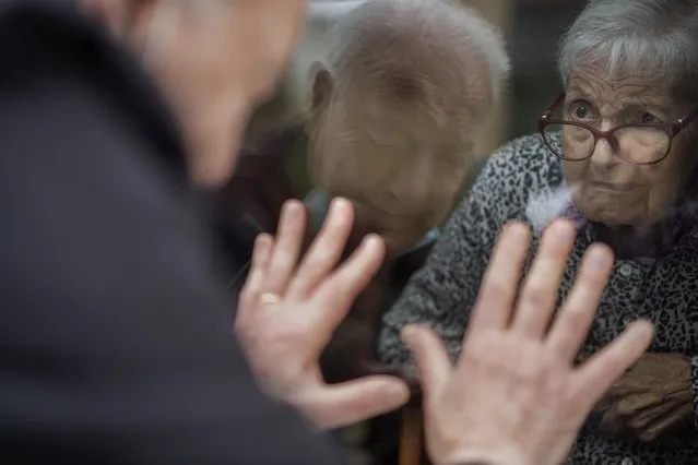 Javier Anto, 90, reacts in front of his wife Carmen Panzano, 92, through the window separating the nursing home from the street in Barcelona, Spain, Wednesday, April 21, 2021. Since the pandemic struck, a glass pane has separated – and united – Javier and Carmen for the first prolonged period of their six-decade marriage. Anto has made coming to the street-level window that looks into the nursing home where his wife, since it was closed to visits when COVID-19 struck Spain last spring. (Photo by Emilio Morenatti/AP Photo)