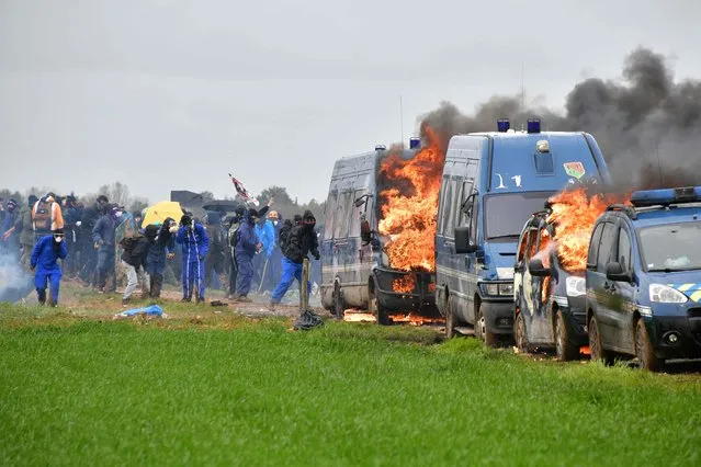 Protestors stand next to French gendarmes' burning cars during a demonstration called by the collective “Bassines non merci”, the environmental movement “Les Soulevements de la Terre” and the French trade union “Confederation paysanne” to protest against the construction of a new water reserve for agricultural irrigation, in Sainte-Soline, central-western France, on March 25, 2023. More than 3,000 police officers and gendarmes have been mobilised and 1,500 “activists” are expected to take part in the demonstration, around Sainte-Soline. The new protest against the “bassines”, a symbol of tensions over access to water, is taking place under thight surveillance on March 25, 2023 in the Deux-Sevres department. (Photo by Pascal Lachenaud/AFP Photo)