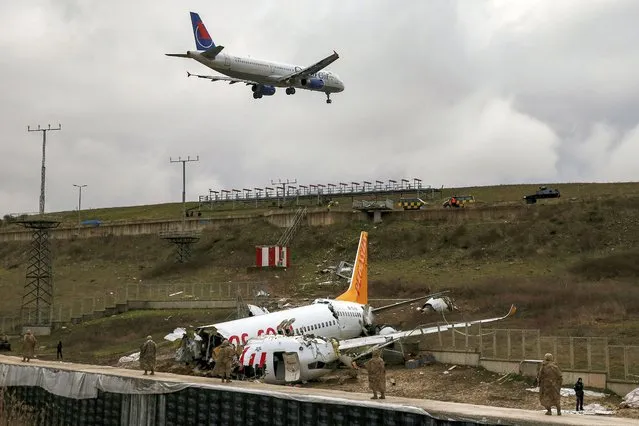 A plane descends to land, as soldiers guard the wreckage of a plane operated by Pegasus Airlines after it skidded Wednesday off the runway at Istanbul's Sabiha Gokcen Airport, in Istanbul, Thursday, February 6, 2020. Flights resumed Thursday after the airliner skidded off a runway, killing three people and injuring dozens. The Boeing 737 landed during strong winds and heavy rain and overshot the runway. It skidded about 50-60 meters (yards) before it dropped into the ditch from a height of about 30 meters (98 feet). (Photo by Emrah Gurel/AP Photo)