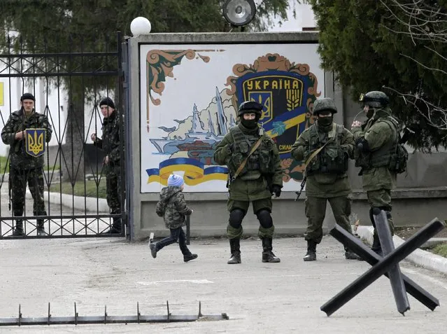 A boy runs by as Ukrainian soldiers look on from behind gates as Russian soldiers guard the gate of an infantry base in Perevalne, Ukraine, Tuesday, March 4, 2014. (Photo by Darko Vojinovic/AP Photo)
